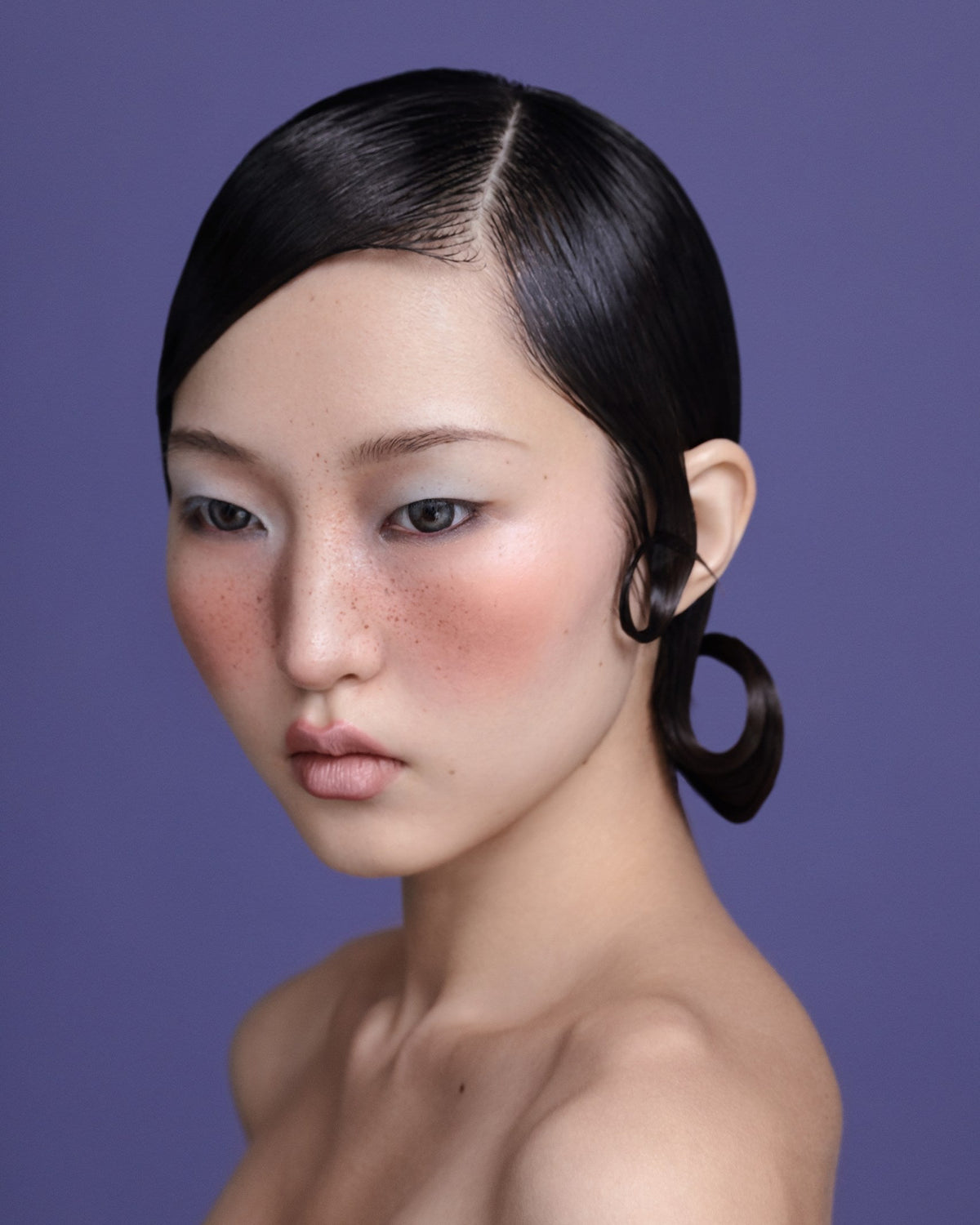 Close-up portrait of a model with sleek, wet-look styled dark hair and artistic curls, showcasing the Ambre hair oil by Yann Beyrie Paris against a purple background.