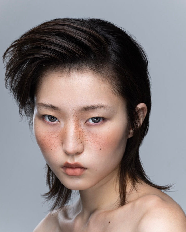 Editorial beauty portrait by Yann Beyrie Paris featuring an Asian woman with a short, textured haircut with subtle mullet styling, natural prominent freckles across her nose and cheeks, and striking grey eyes against a neutral grey background.