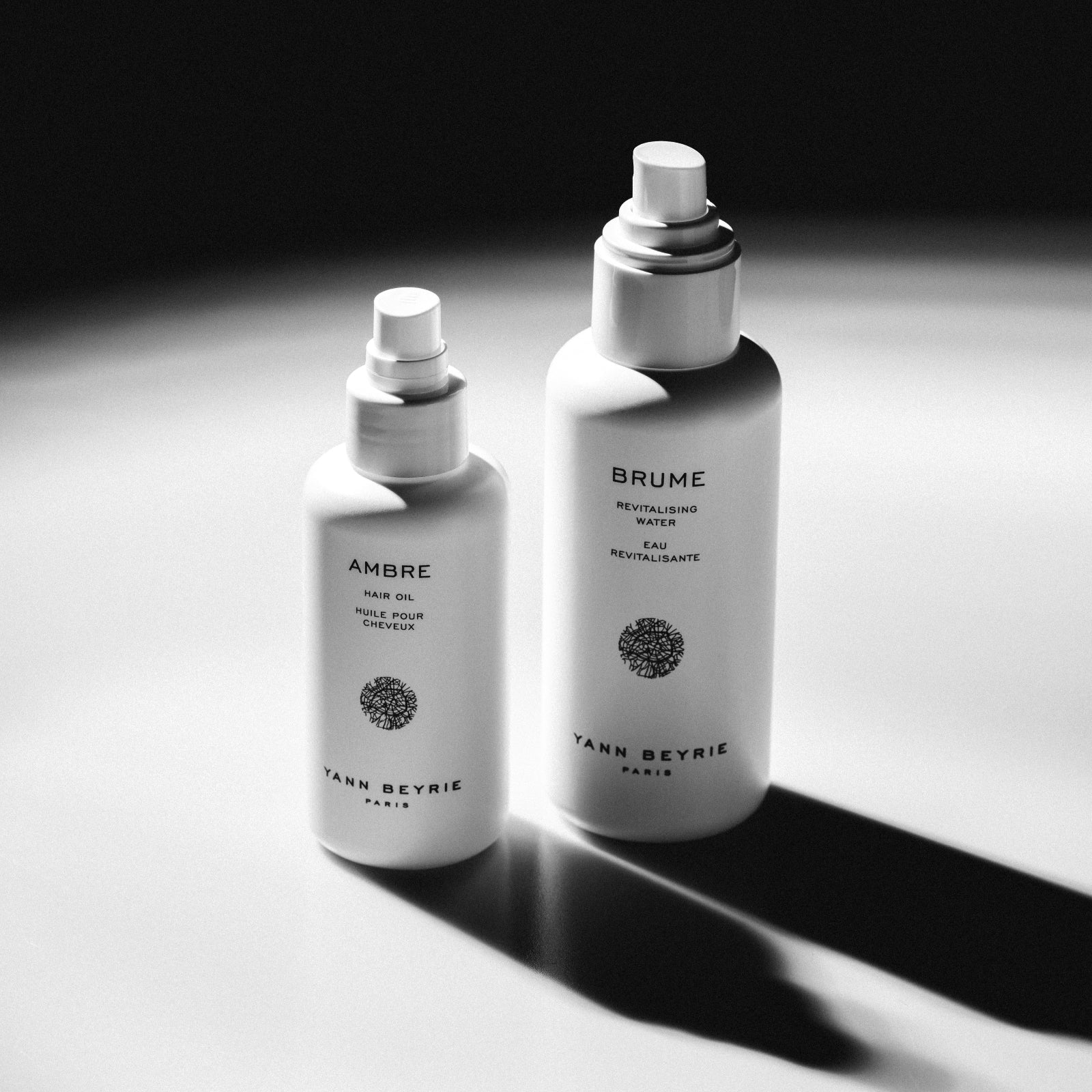 Minimalist black and white studio shot of Ambre Hair Oil and Brume Revitalising Water bottles with dramatic shadows