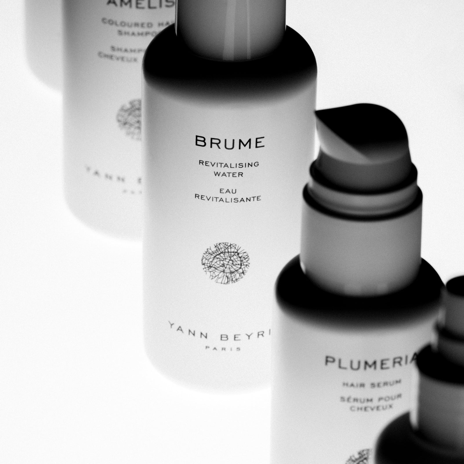 Close-up black and white photo of Brume Revitalising Water and Plumeria Hair Serum bottles by Yann Beyrie Paris