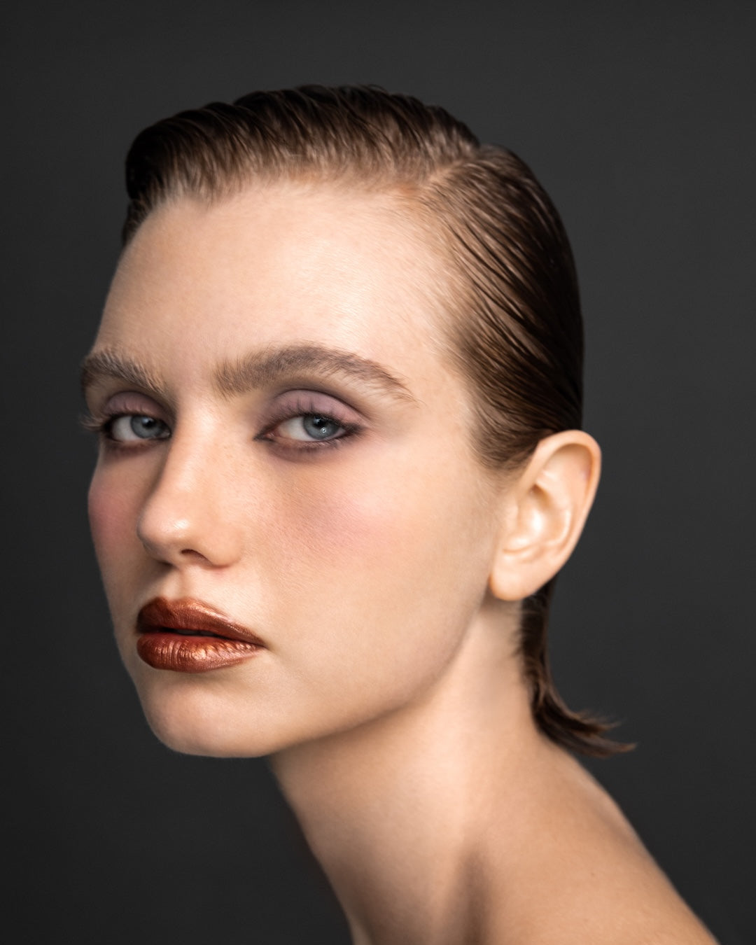 Female model with sleek slicked-back hair, purple eyeshadow, and copper metallic lips at Yann Beyrie Paris