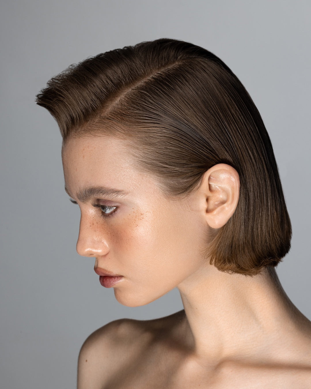 A professional profile portrait of a model with a smooth, tailored medium-length brunette haircut, showcasing vibrant hair color and mirror-like shine, promoting the Amelis Colour Care Shampoo by Yann Beyrie Paris against a soft grey background.