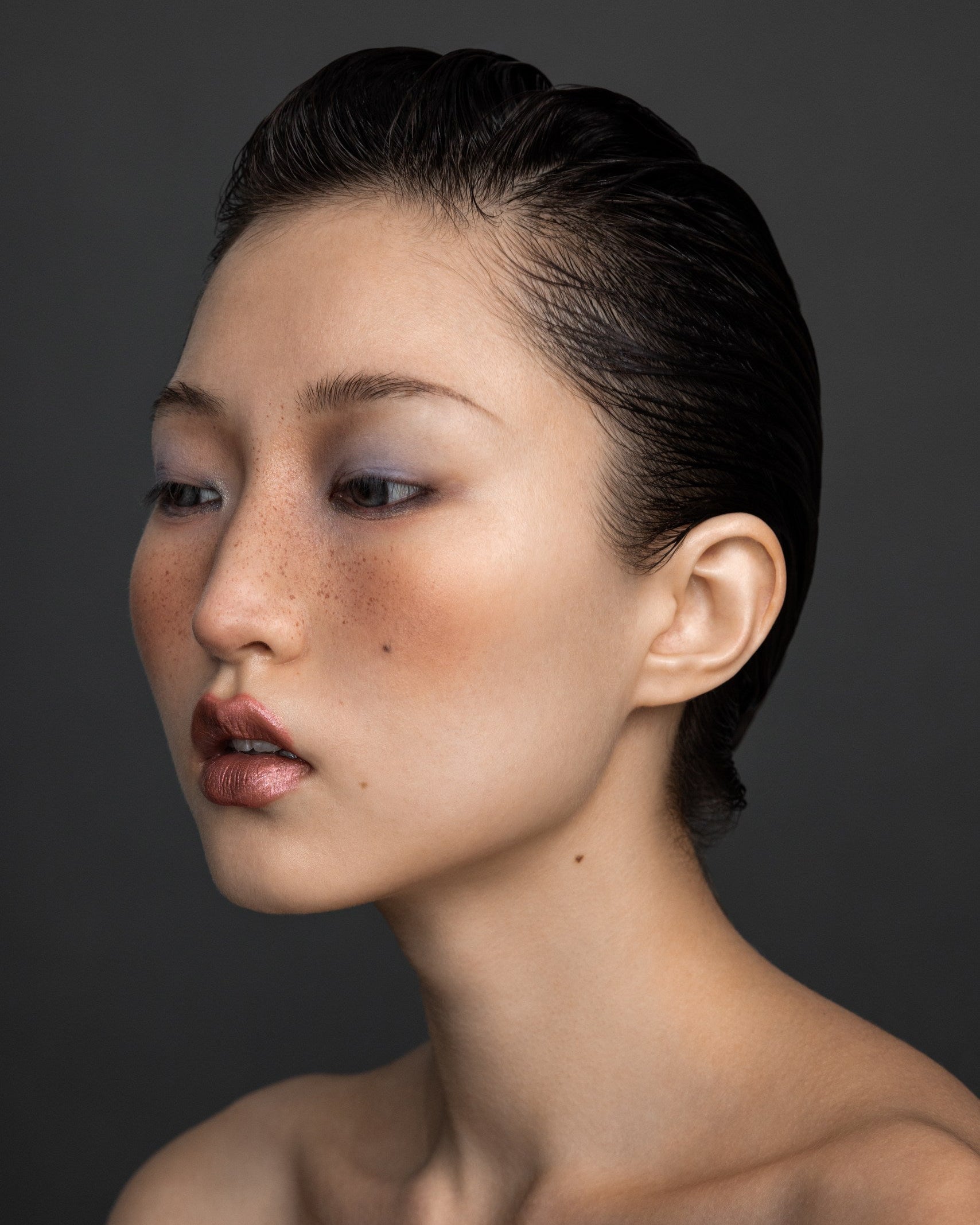 Close-up profile portrait of a model with a sleek, wet-look hairstyle, showcasing the high-gloss finish and frizz control achieved with Plumeria Hair Serum by Yann Beyrie Paris against a dark grey background.